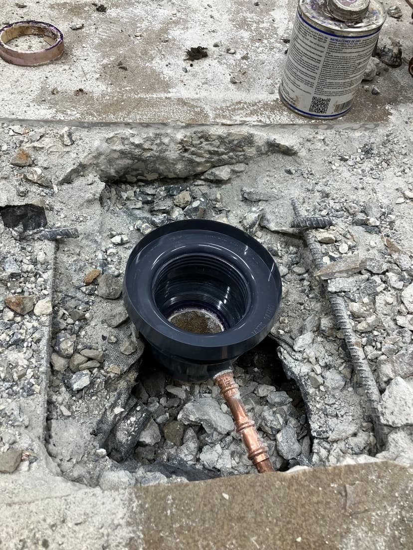 Pipe fitting installed in a concrete floor, surrounded by broken concrete and debris.